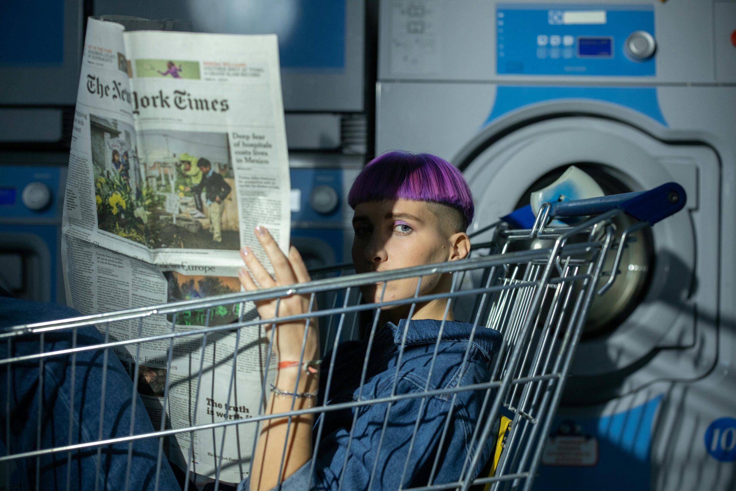 A young individual with purple hair reads a newspaper in a laundromat, sitting in a shopping cart.